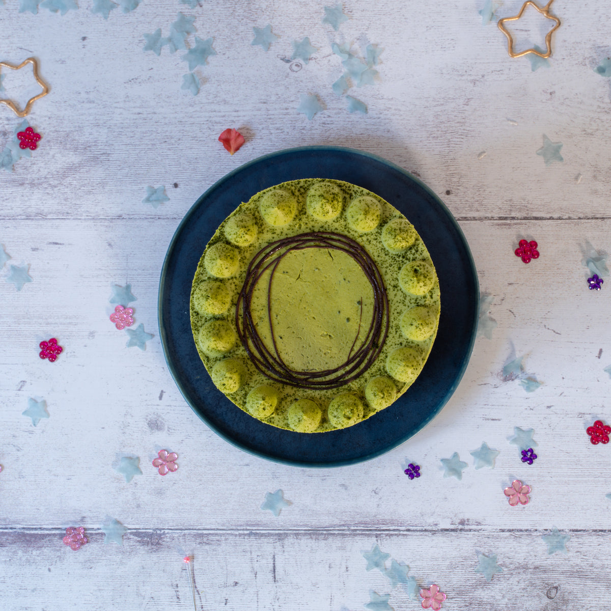 Green Matacha cake with chocolate drizzle on a blue plate against a wooden background with decorative stars and flowers.